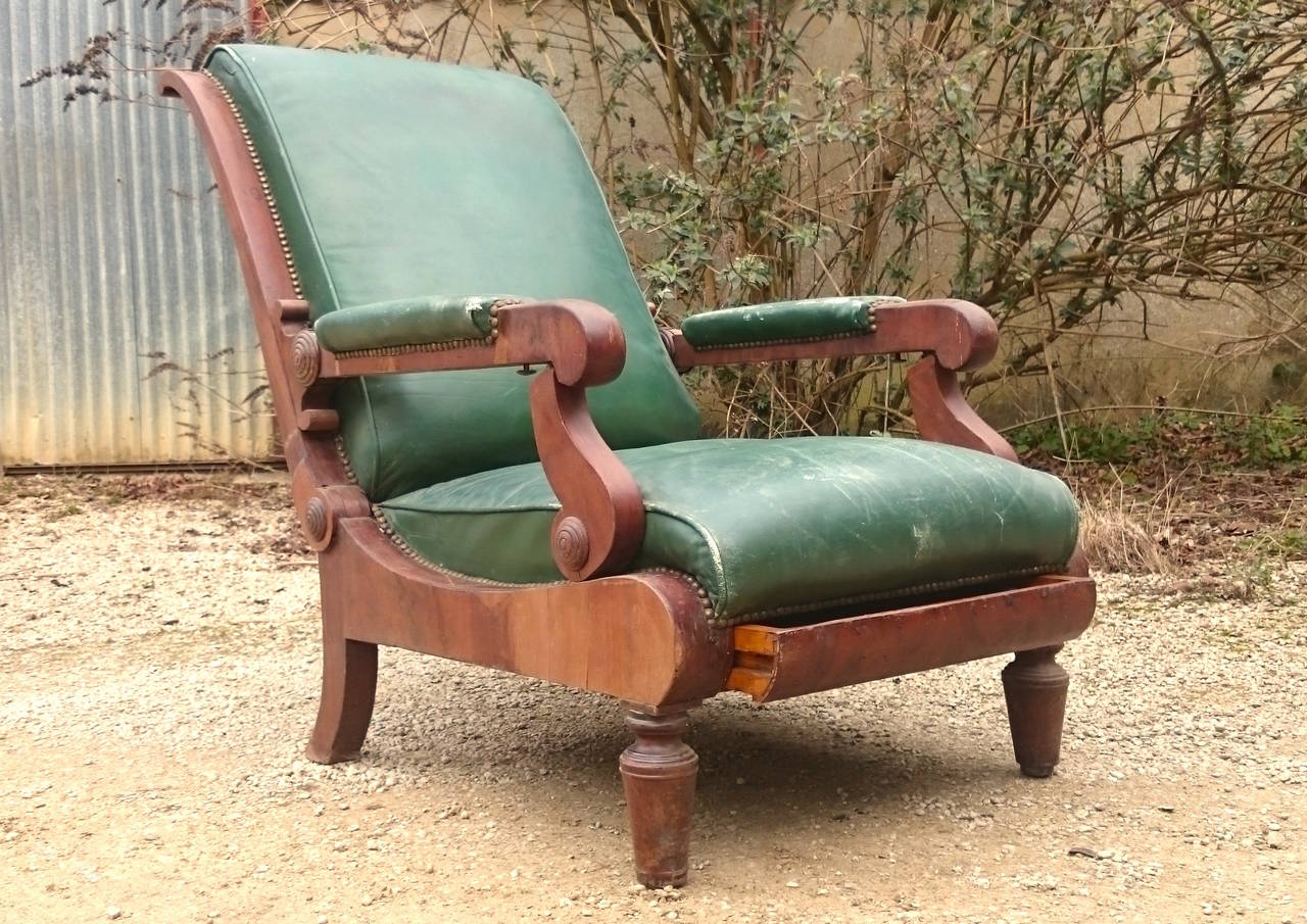 Antique reclining library chair with built in foot rest covered in green leather. The show wood is veneered with flame mahogany. The mechanism is actuated by two buttons hidden underneath the armrest and the foot rest springs up when the drawer is