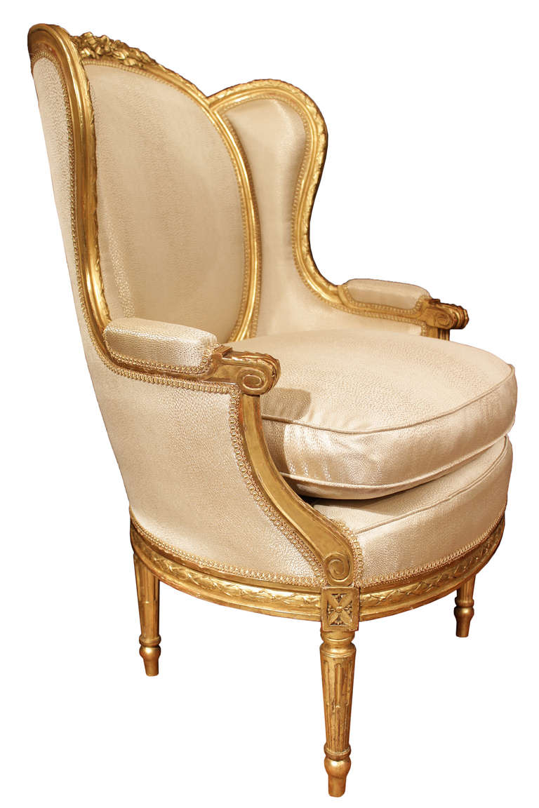 A stunning pair of French 19th century Louis XVI st. giltwood Bergères à Oreilles. Each  wing chair is raised on tapered reeded supports with chandelles and topie shaped feet. The impressive finely carved apron with berried laurel garland border