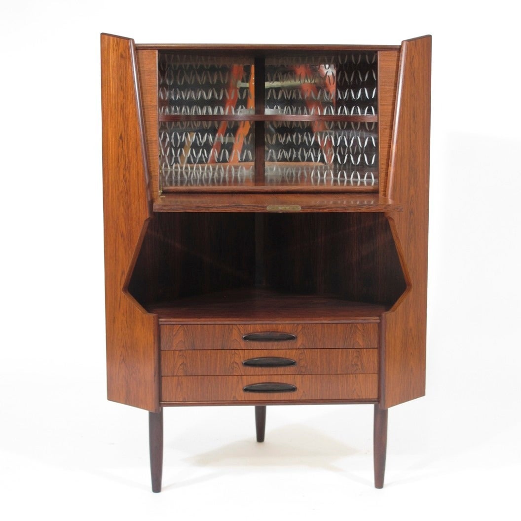 Mid-century Brazilian rosewood corner cabinet with with rich book-matched grain patterns. Drop front Locking door reveal an interior bar with mirrored back with etched pattern; open space and three drawers below. Raised on round tapered legs.