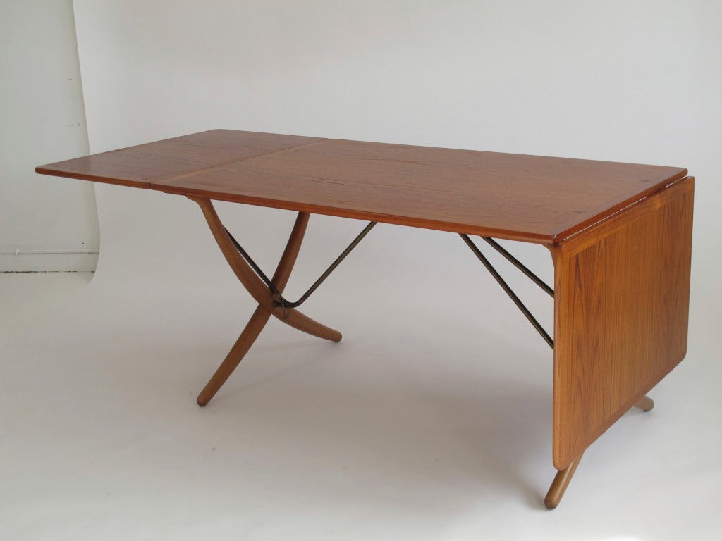 Large drop leaf dining table designed by Hans Wegner for Andreas Tuck Denmark. Teak table top with solid edge trim and two drop leaves. Brass cross bars and support mechanism. Oak curved X shaped legs. 52