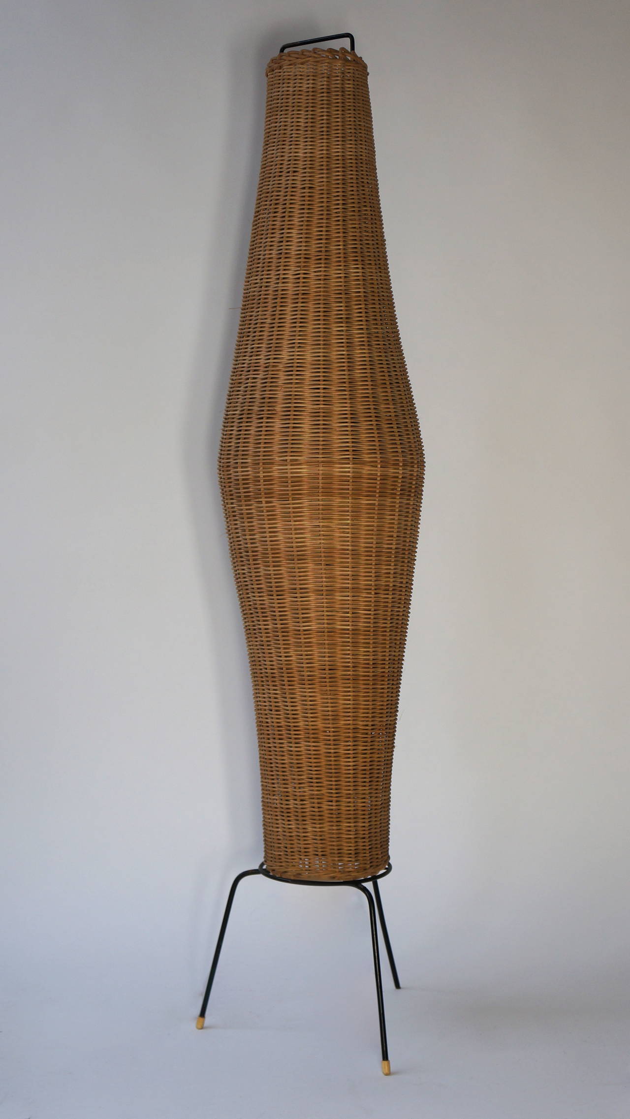 A large-scale floor lamp with a woven wicker body in an asymmetrical 'fishing basket' form with a handle at the top all supported by a wrought iron tripod base.