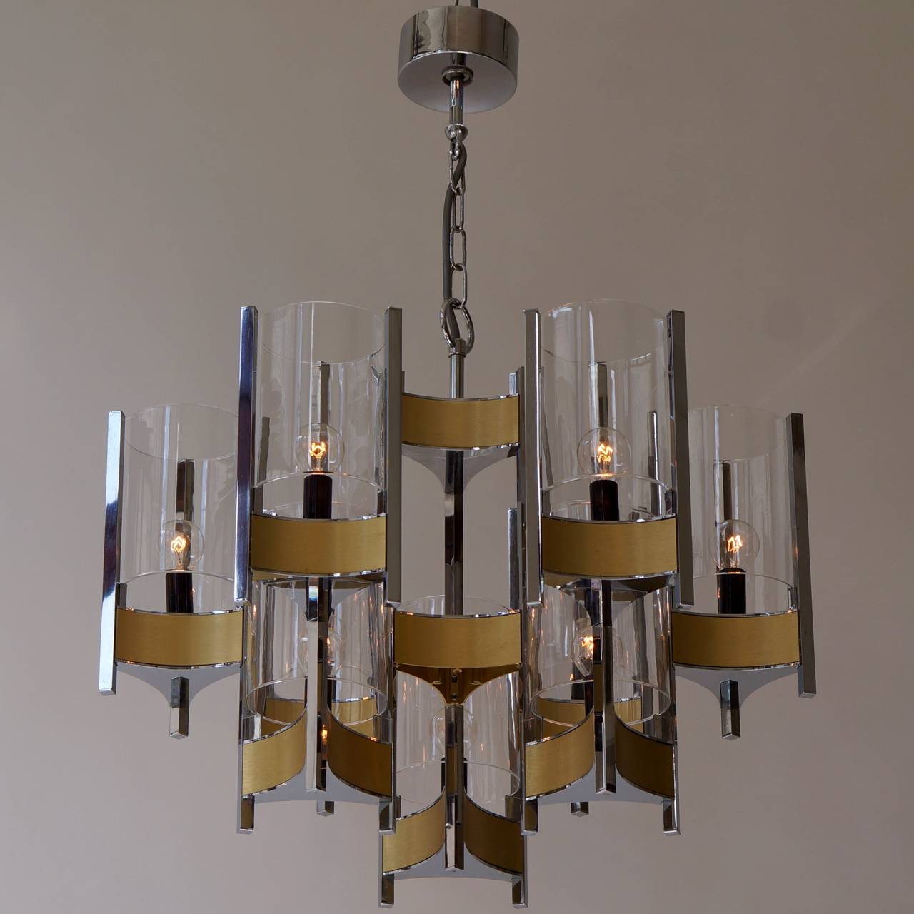 Four Gaetano Sciolari chrome and brushed brass two-tier nine-light chandeliers with cylindrical glass shades, stylish monumental Sciolari chandelier. 

Consist of three arms with glass cylinders. Chrome and brushed brass base.  

We can supply, free