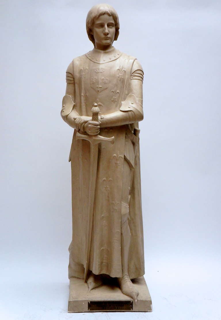 A life size plaster sculpture representing Jeanne of d'Arc as an androgynous girl dressed as a knight and wearing a fleur-de-lys covered cloak, holding a long sword in her hand.
Sculpture by Andre Besqueut (French sculptor born 1850), presented at