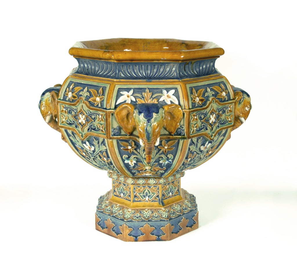 Stoneware with multi-colored glazes. The urn and pedestal modeled in a Hispano-Moresque style with panels of flowers and foliage interspaced with elephant masks, in shades of sand, blue and green.   

This fine example of Faience was created by