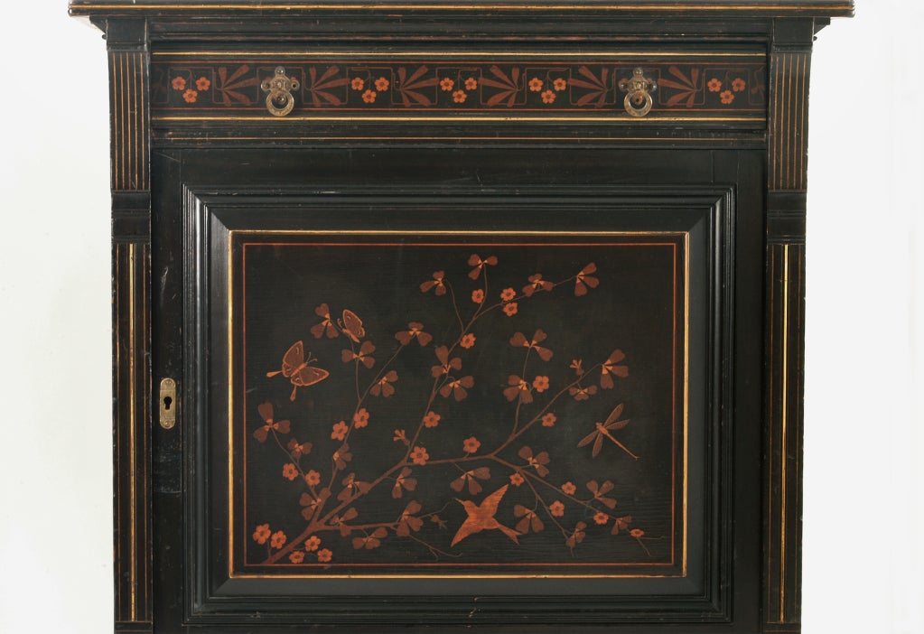 Ebonized cherry wood with elaborate inlaid marquetry of lighter woods as well as parcel gilding and an inset velvet top. Brass hardware. Having a central marquetry door with an inlaid frieze drawer above. The whole resting on Asian-inspired canted
