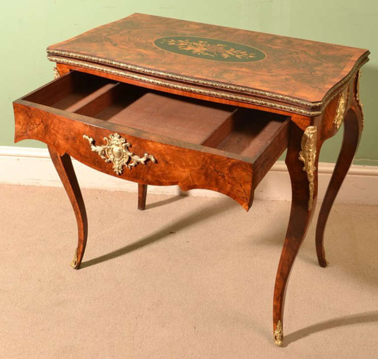This is a lovely antique Victorian burr walnut and marquetry folding games table, circa 1870 in date.

The serpentine fronted and tulipwood crossbanded top has an oval central floral marquetry panel. The hinged top opens to a baized interior and