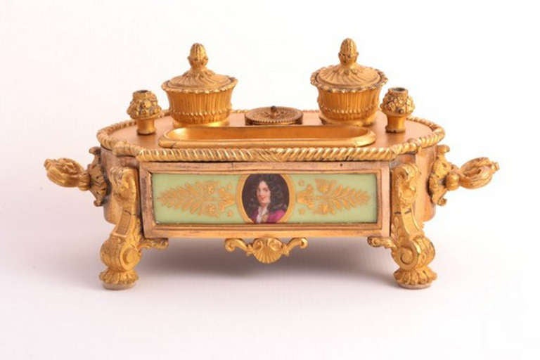This is an antique French ormolu and porcelain encrier of rectangular form c.1870 in date. 

The inkstand compromises of two inkwells centered by a pounce pot and a pen tray. The inkstand is further decorated with two hand painted porcelain