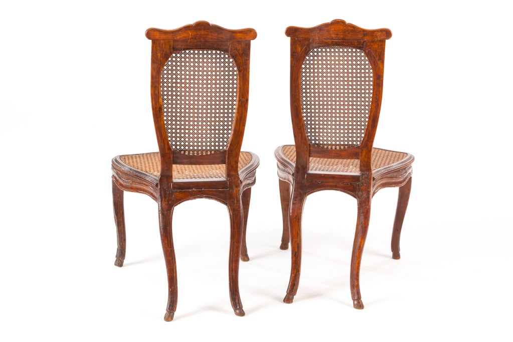 Carved wooden chairs, slightly tilted backs with cane seats, for women to sit on allowing for their pannier skirts.