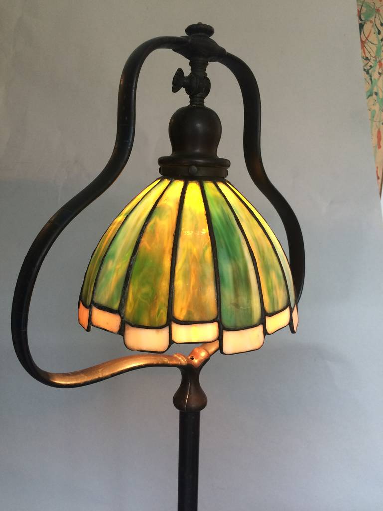 Cast bronze floor lamp with signed, original shade by Handel. Circa 1910. Bell shaped leaded glass shade refracts a soft glow, with playful white sawtooth edging. Articulating knuckle joint allows light to tip to the right or left for armchair