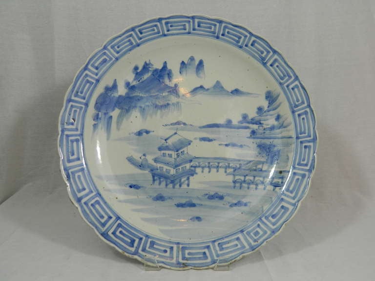 19th century large Japanese Arita charger, the wide scalped-edge with key fret band framing the under glaze blue painted landscape with a lakeside viewing pavilion.