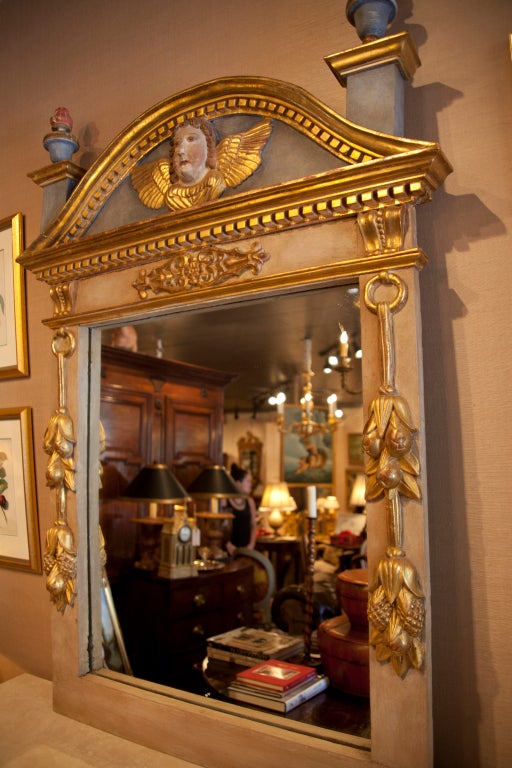 Miroir en verre de style néoclassique, polychromé et doré à la feuille, 19e siècle. La plaque rectangulaire est surmontée d'une crête bombée et dentelée centrée par un masque ailé, flanquée de part et d'autre d'un fleuron en forme d'urne flamboyante