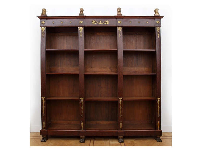 This bookcase in clouded mahogany has three vertical divisions. The architrave and four pilaster-like stiles are embellished with classically-inspired gilt bronze ornaments, including griffins, palmettes, garlands, floral motifs, stars, birds and