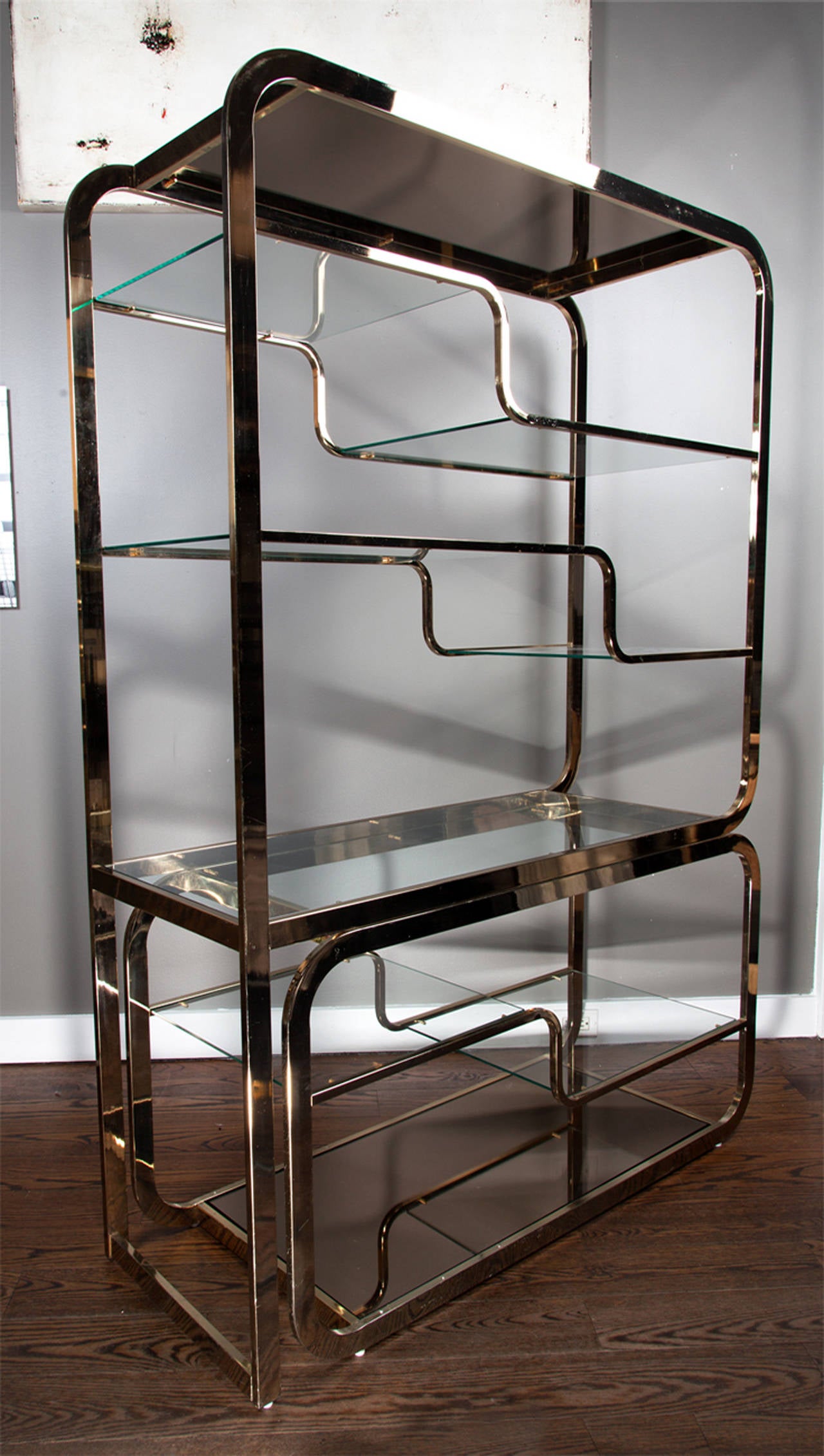A brass Milo Baughman étagère with glass shelves. The two-piece design allows the bottom tier to be extended to the side, allowing for additional storage. The bottom shelf has a bronze inset mirrored top; the rest of the shelving is clear glass.