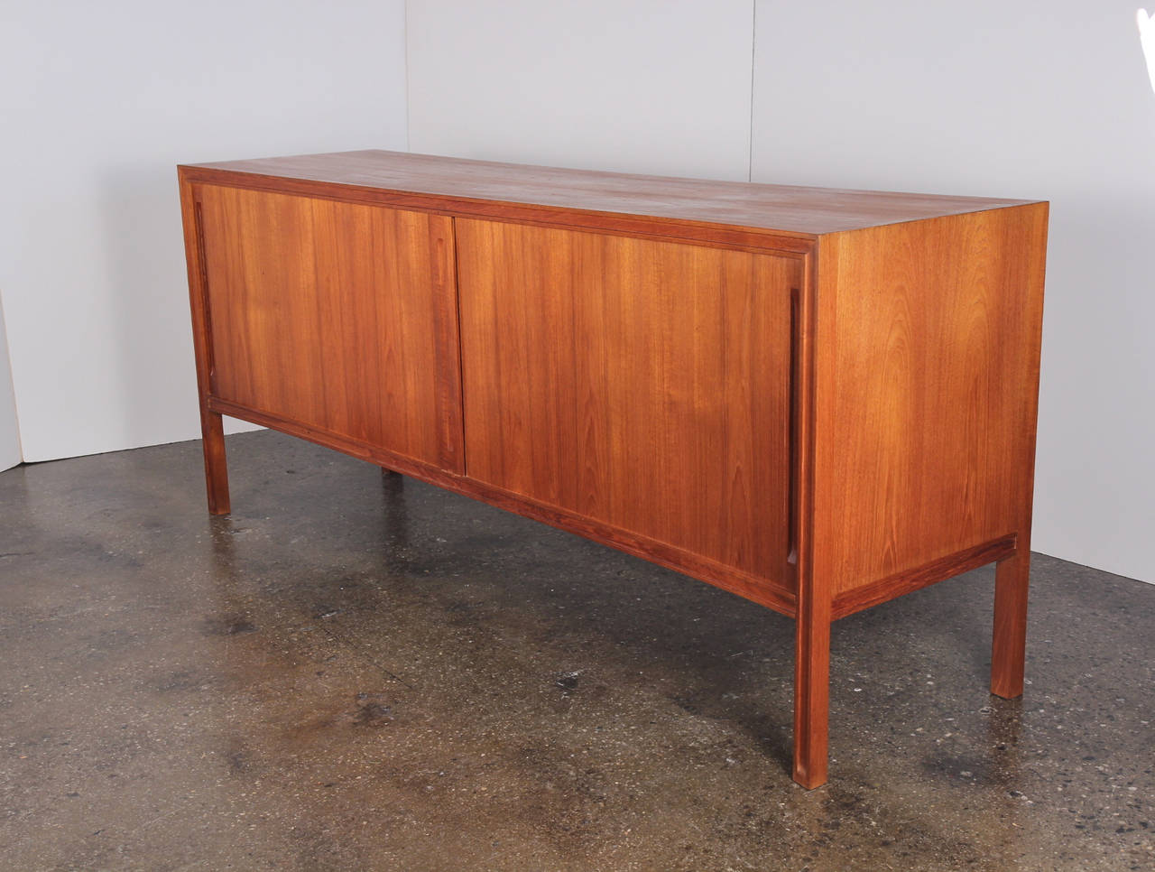 Tambour doors and a curved front create understated elegance in a minimalist teak sideboard. Plenty of storage with seven drawers and adjustable shelves. Mitered trim frames its face and subtle channels decorate the legs. A finished back means it