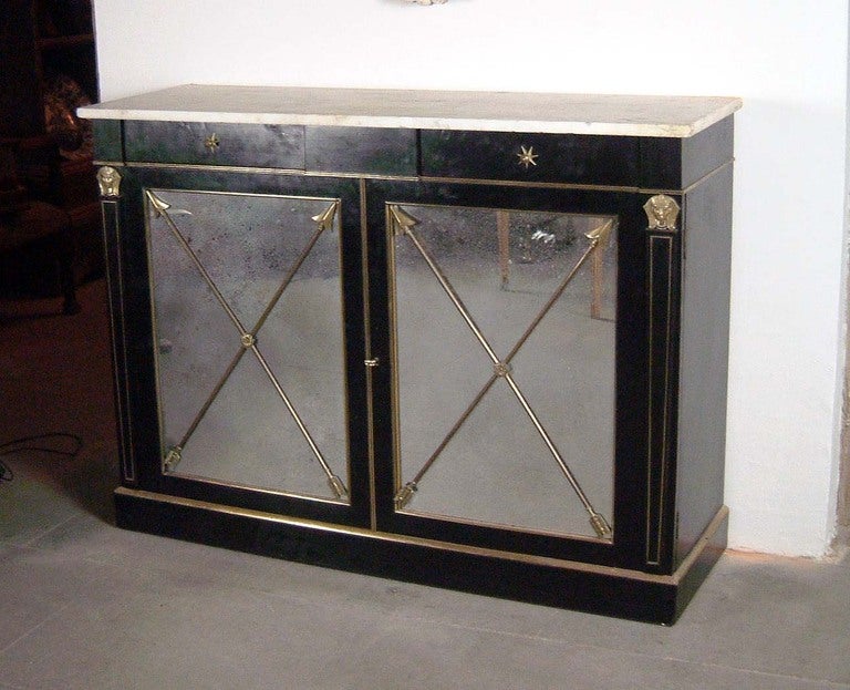 A black lacquered wood cabinet with two mirror doors fitted with brass mounts.
Two metal arrows crossing each mirror.
Motted white marble top.
Two front drawers with metal tiroirs.
Shelves inside.
