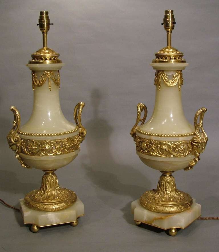 Converted into lamps from their original vase forms, and constructed in the Louis XVIth style, using Algerian onyx and gilt bronze; the square form incurved angle bases supported by gilt bronze ball feet; the waisted bronze socles holding baluster
