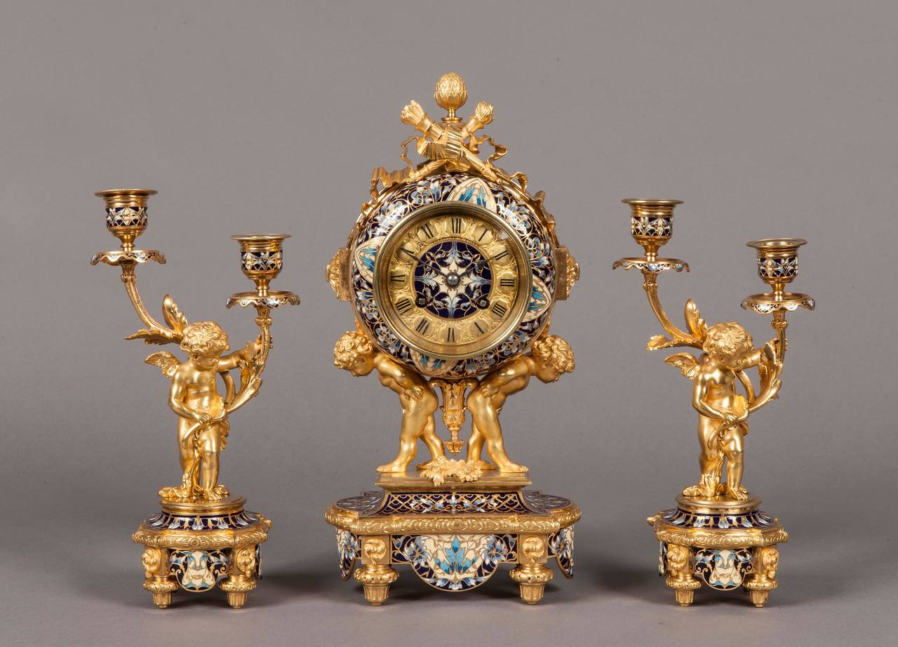 Constructed in gilt bronze and decorated with champleve´ enamel; the candelabra have toupie feet supporting the shaped circular bases, on which winged cherubs hold two armed candle arms aloft; the shaped elliptical form clock has conforming feet,
