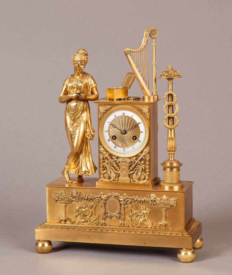 Constructed in Gilt Bronze, and rising from toupie feet, the rectangular base exuberantly decorated with applied designs and motifs drawn from the source books of Messrs Percier and Fontaine; the clock case surmounted by a Grecian lady examining a