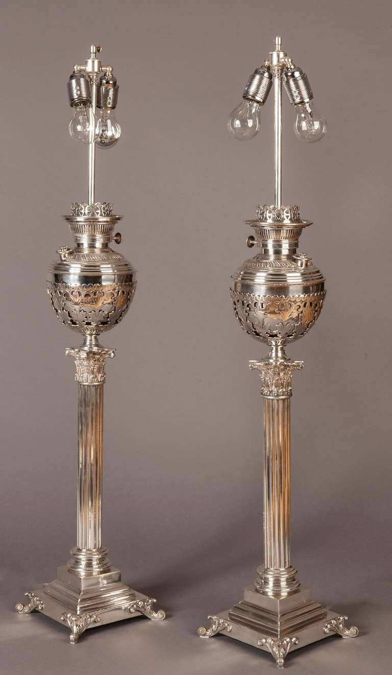 Silver plated, designed in the Neo-classic manner, the square stepped bases supported by claw feet, the circular central columns terminating with Corinthian capitols, and ovoid oil reservoirs complete with the burners, encased within pierced