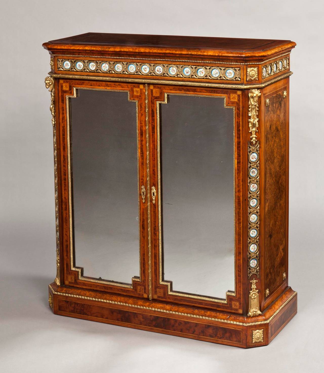A Fine Pair of Side Cabinets

Constructed in Circassian walnut and various specimen woods, exuberantly mounted with porcelain plaques and quality gilt bronze mounts; rising from plinth bases, having canted angles, dressed with rams head spandrels