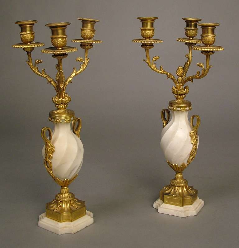 A good pair of candelabra in the Louis XVI manner.

Constructed in Carrara marble and gilt bronze; the square stepped bases having incurved angles, and supporting bronze socles dressed with pendant bellflowers, holding baluster form urns decorated