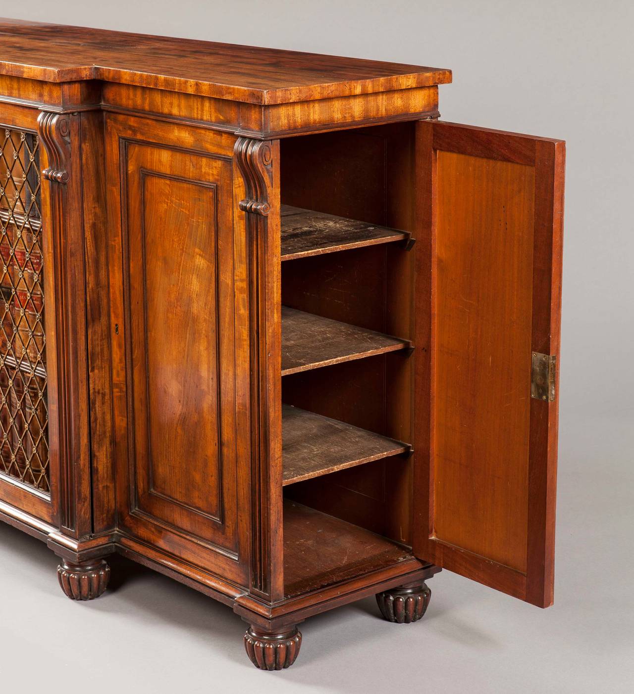 Of four door breakfront form, and constructed in a fine Honduras mahogany, rising from carved oblate feet; set within upright reeded stiles terminating in spandrels, the two lockable centre doors are dressed with brass trellis grills, having raised