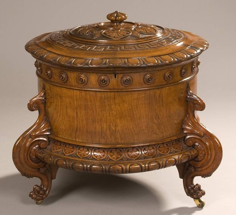 A highly decorative wine cooler in the Graeco-Roman taste 

Of cylindrical form, constructed in well figured oak, supported by tripartite feet, of complex winged foot form, with brass castors under: the body with a lobed lower rim, with a running