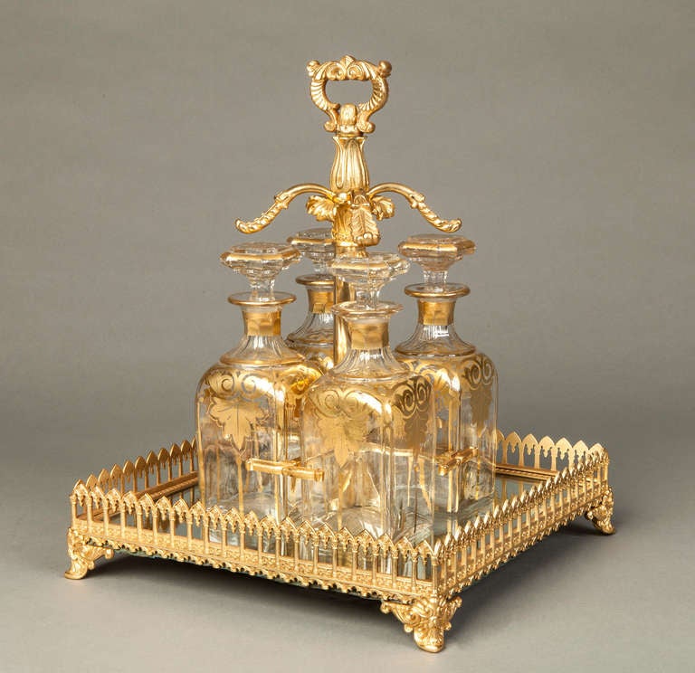 A drinks set of the Napoleon III Period

The ormolu rectangular frame, rising from swept feet, with a running arcaded gallery, fitted with a mirror plate base houses a suite of four lead glass crystal stoppered decanters of square form, with