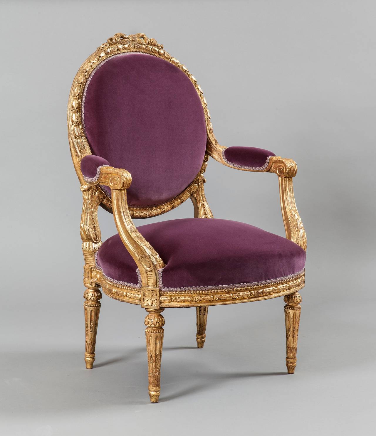 Constructed in a well carved gold giltwood, with later purple velvet upholstery; rising from turned and tapering legs with toupie feet, the shaped front rails embellished with a carved running Bellflower band; the backs of elliptical form, with