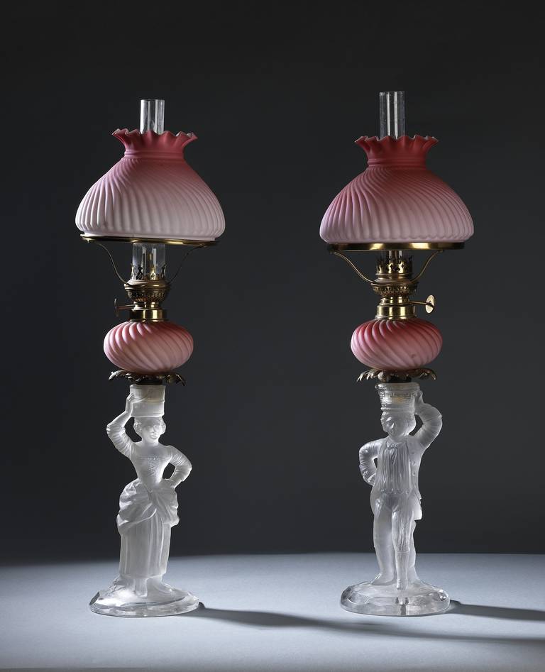 Supported by opalescent glass figures of a young man and lady in Victorian attire; the oil reservoirs and shaped shades in ‘peach blow’ Stourbridge glass, fitted with the patent ‘Kosmos Brenner’ wick and fuel mechanism.