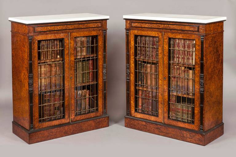 Constructed in burr yew, with ebony contrasts, bronze trellis work grills and carrara marble; rising from plinth bases of rectangular form, with addorsed gadrooning over; the lockable double doors fitted with trellising enclose shelved interiors;