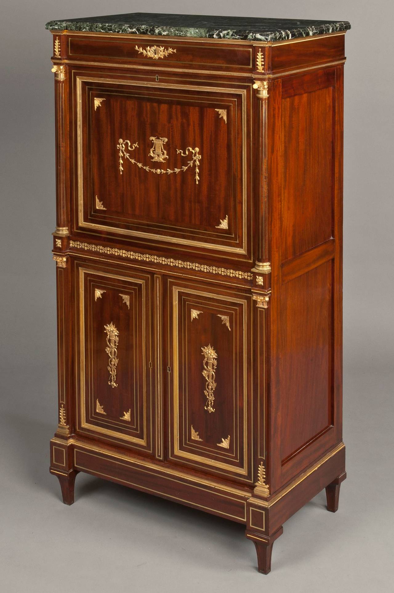 The case constructed in mahogany, with gilt bronze accents; rising from inverted truncated pyramidical legs; the platform of Verte de Mer marble with a thumbnail moulded edge, a long drawer below, with the fielded panelled fall front having running