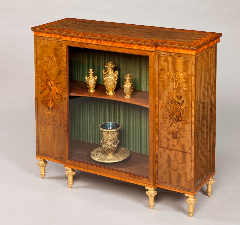 Constructed in a beautifully figured satinwood, with finely executed marquetry inlays; of restrained breakfront form, rising from gilt toupie feet, turned and dressed with ‘running pearl’ annuli, the shelved central open display sections, having