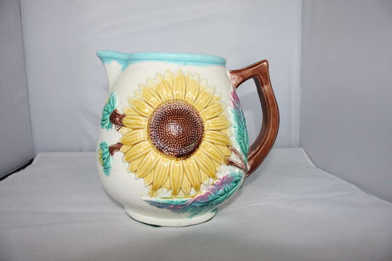 Antique pitcher with large sunflower motif on one side and yellow lily on the other.  Pitcher has brown stem like handle and shell pink interior and green leaves.