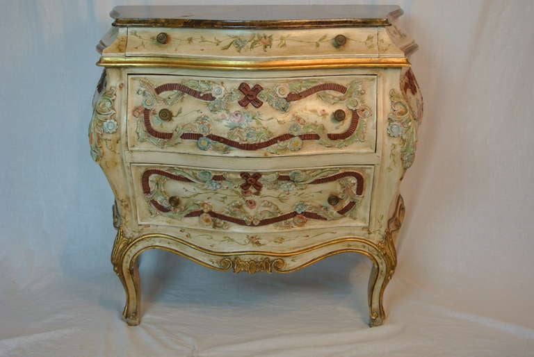 From Howard Kaplan, NYC, this chest of drawers has accentuated curved sides and front. Deeply carved all around in floral and ribbon motif. Antiqued cream ground with poly chrome details. 3 drawers.