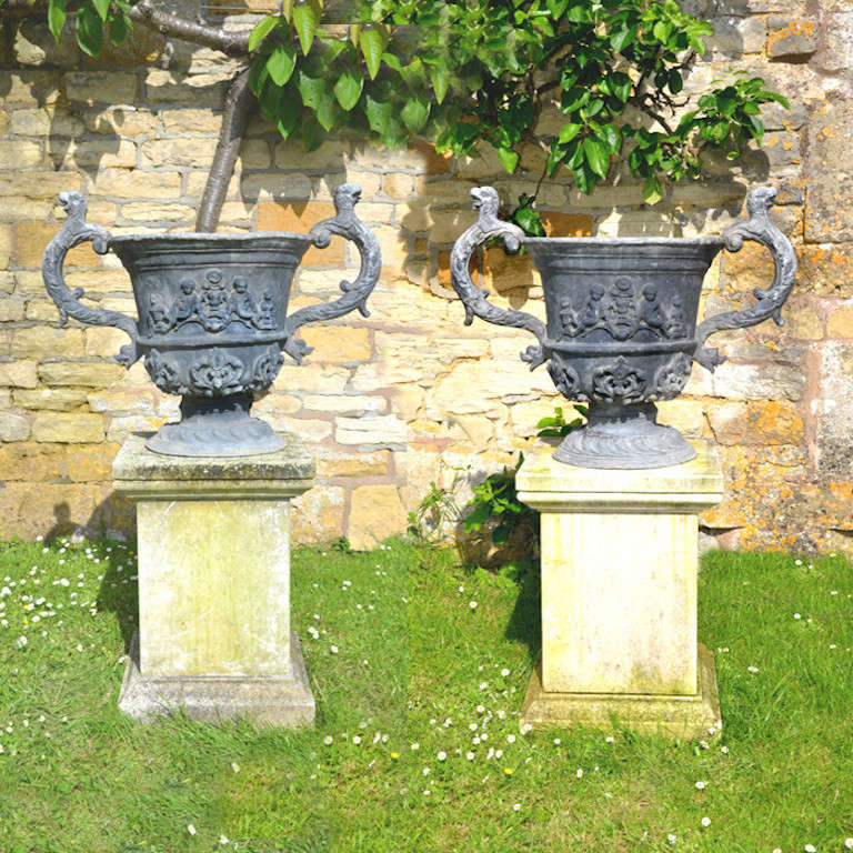A pair of large early 20th century lead urns embellished with fleur de lys and cherubin decoration, surmounted by griffin and dolphin handles, raised upon a circular socle.