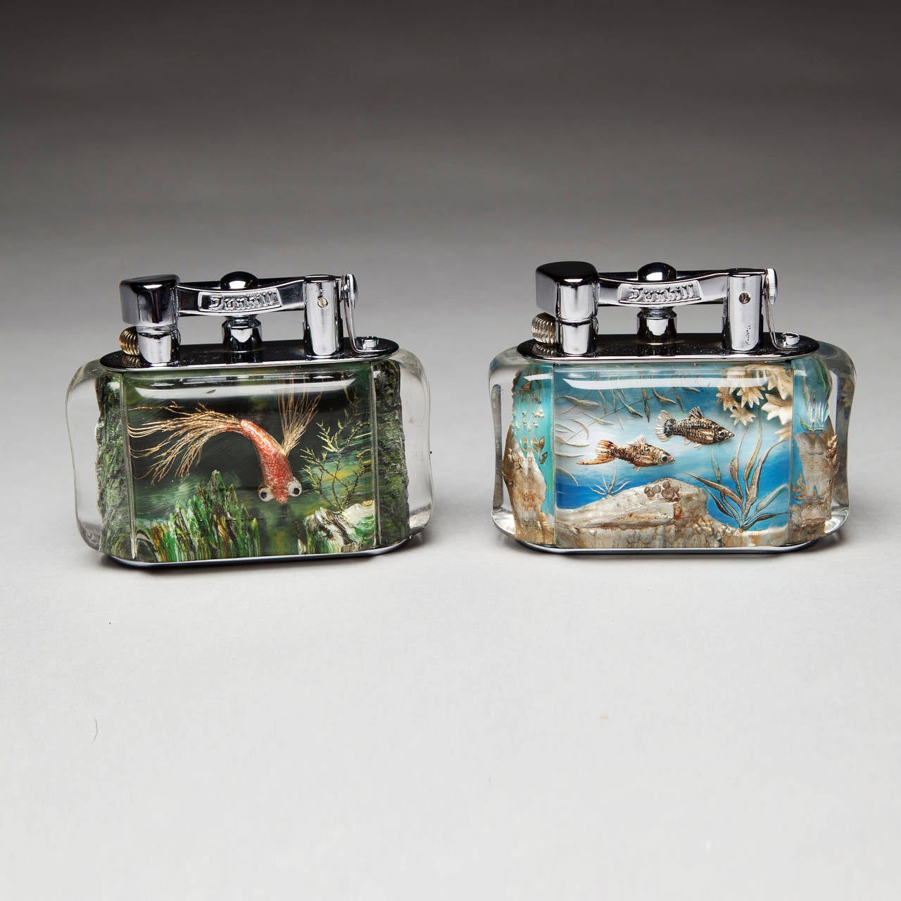 Two very bright and well preserved chrome plated Dunhill Aquarium Table lighters, the lucite panels boldly cut and reverse painted with well studied fresh water fish among reeds and rocks.   

In perfect working order.

England, circa 1950-60