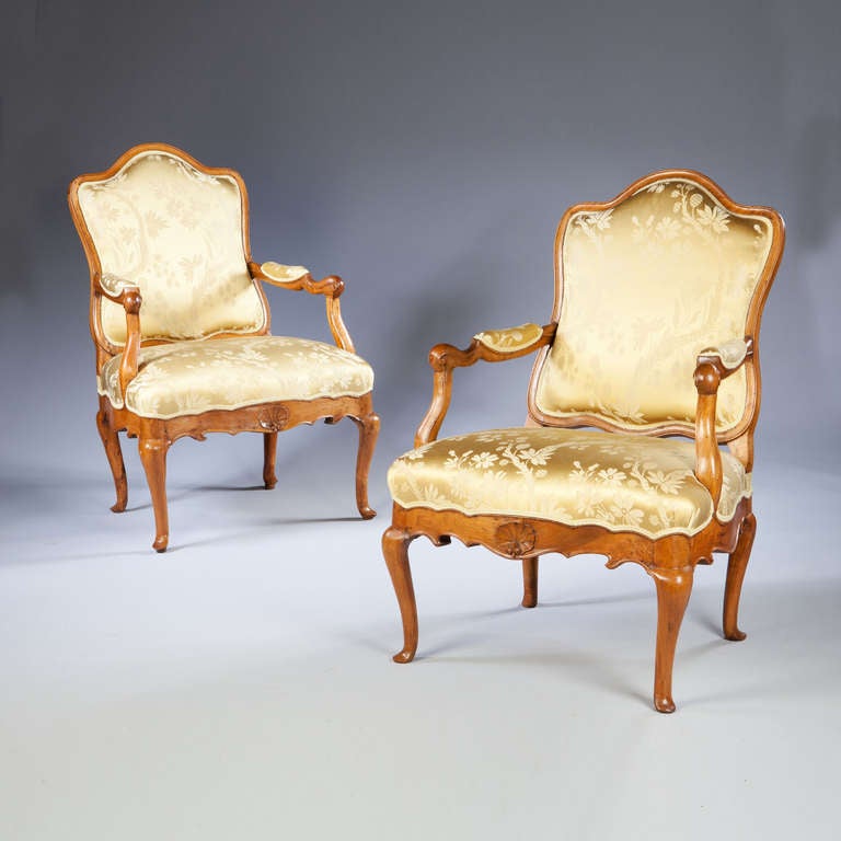 A pair of Genoese walnut fauteuils with scrolling curvaceous backs and scroll arms, serpentine rails. The front carved with a shell, standing on cabriole legs.