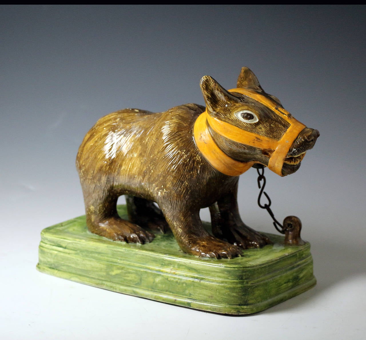Rare unrecorded figure of a bear cub modeled muzzled and tethered with a metal chain on a green base. The figure is a significant size and is strongly decorated in pratt colours. 
Bear baiting was a popular 