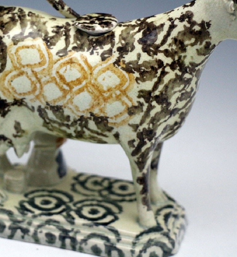 An unusually decorated pottery cow creamer from the early 19th century. 
The figure is modelled standing on a base with a rather diminutive milk maid at work. 
The sponge decoration is enhanced by a diamond shaped patterns on the body and circular