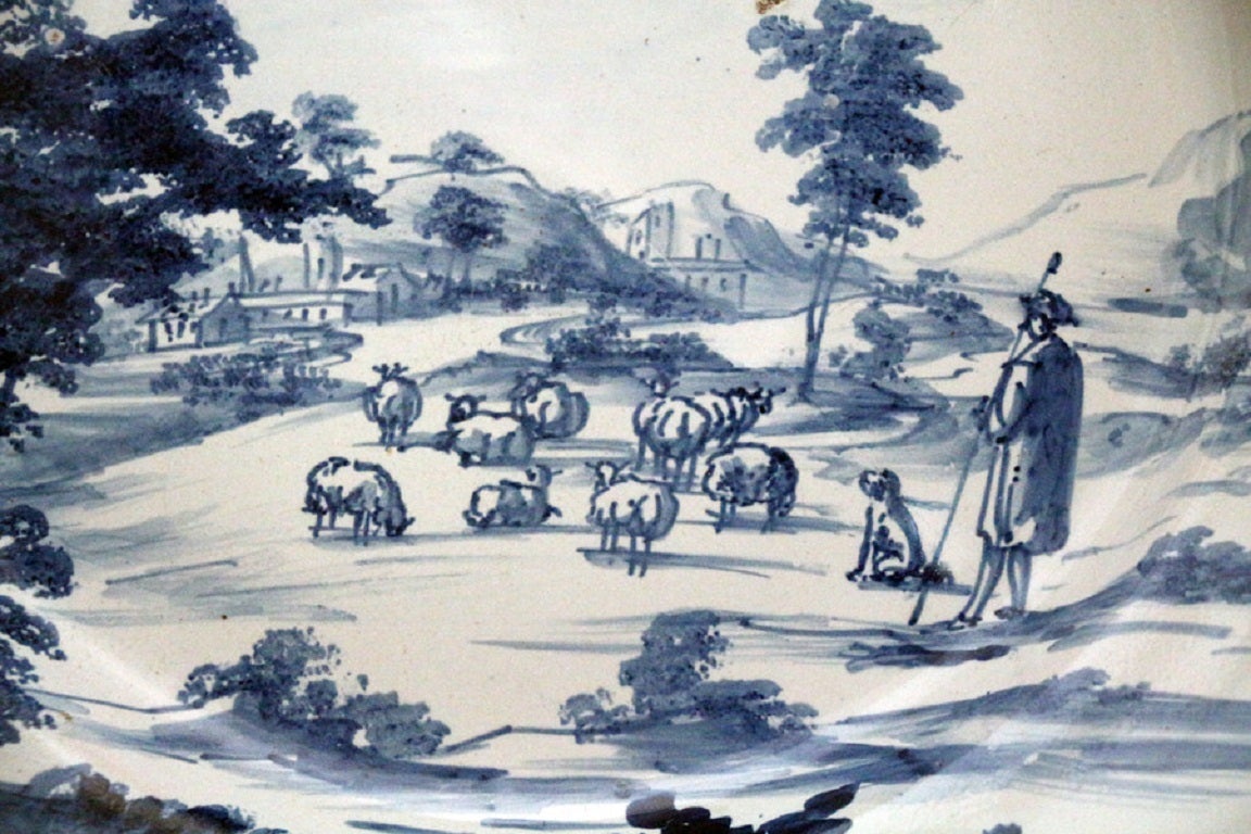 English earthenware blue and white delft charger, a romantic rural scene with a shepherd and his flock with mountains in the background. 
Mid 18th century.