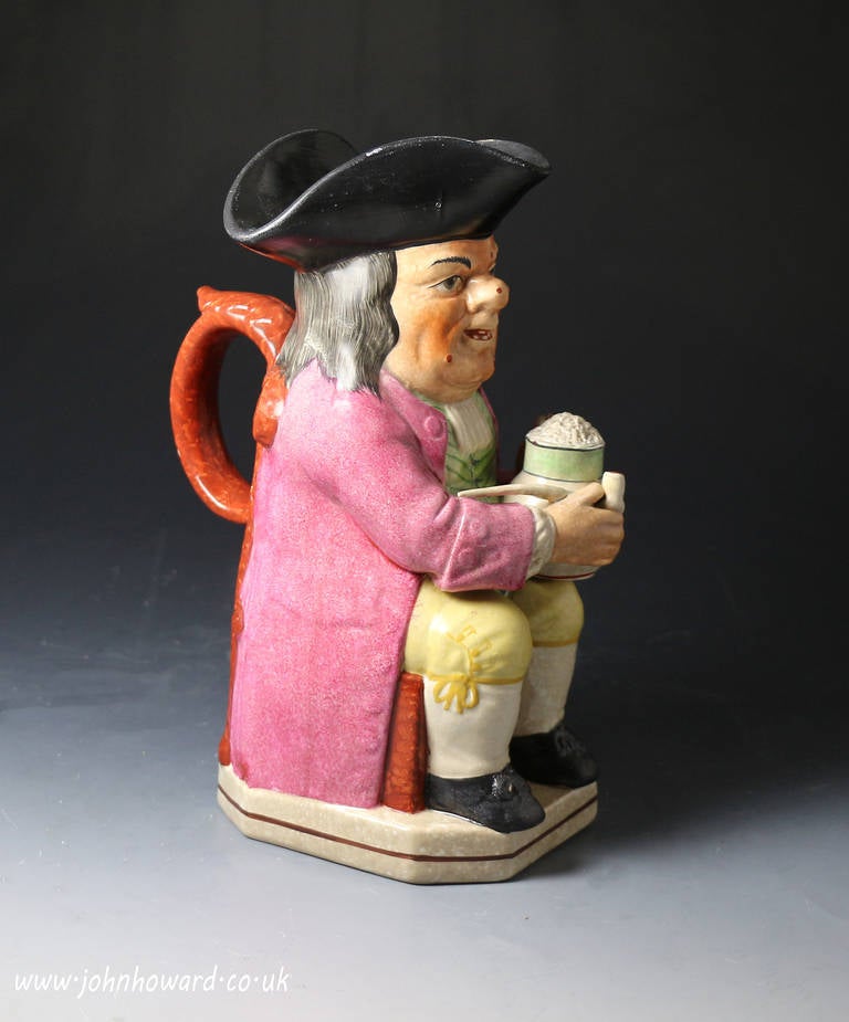 Staffordshire Pottery pearlware glazed Toby Jug. 
The figure is wearing a long coat and tricorn hat whilst seated with a frothing jug of ale and tobacco pipe in the other. 
Depite his facial spots and warts the character is enjoying his