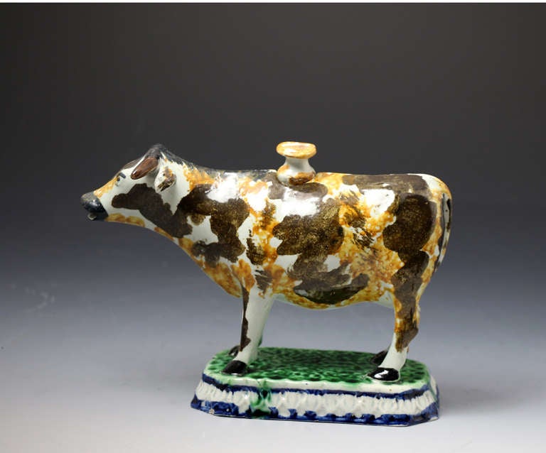 Antique pottery cow creamer in the Prattware color palette. 
The figure of the cow is exceptionally well decorated and is modelled standing on a vibrant verdant green base with cobalt blue borders. The original cover of the figure is an unusual and