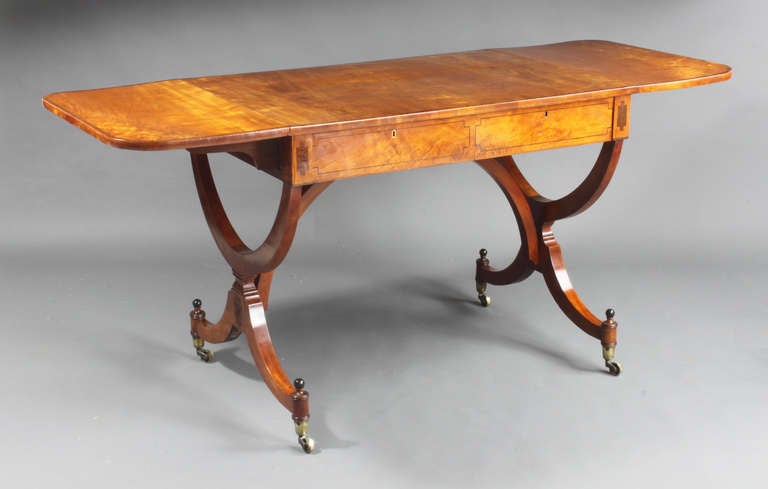 A satinwood sofa table with lovely matched veneers and tulipwood cross banding on the top; the drawers, which are designed without handles, have ebony stringing and the standard end bases also have ebony balls above the castors and carved paterae on