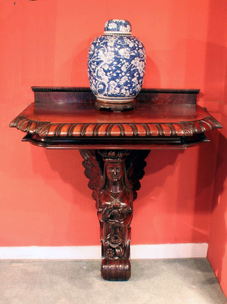 An angel console table in mahogany. The winged angel is wearing a crown of leaves and fruit and holding flowers and fruit in her hands; the table has a bold dragooned edge and a small 3