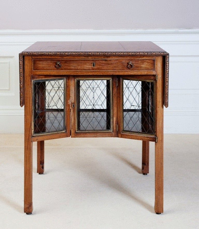 A George III padouk breakfast table after a design by Thomas Chippendale, the rectangular twin flap top with a floral carved border above a frieze drawer and a shelf with brass framed wire sides and a double hinged door on square chamfered legs.