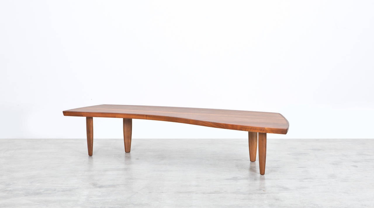 The handcrafted coffee table comes with a walnut veneer top and is supported by four tapered dowel legs. The table is designed by famous American George Nakashima and can also be used as a bench. Another example in stock. Manufactured by Widdicomb.