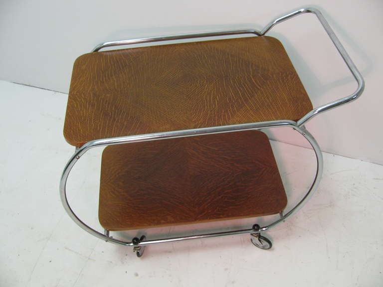 Streamlined mid century bar cart influenced heavily in its design by Bauhaus Modernism. Nickel tubing surrounds the top of quarter sawn oak in geometric panels. Round bakelite locking hardware secure wheels to the frame. In excellent vintage