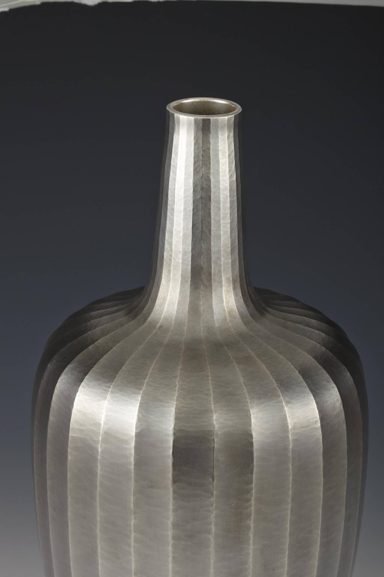 Vase in a cylindrical, bottle-form, the sides finished in thin, vertical facets. Of uchidashi or hammered silver. Marked on the reverse: Jungin or Pure Silver and signed with a chiseled signature by the artist: Juko and with a kao (Hirata Juko II,