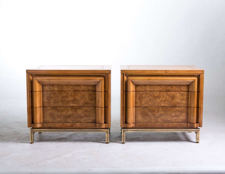 A pair of Mastercraft style three-drawer chests with burled wood fronts and brass stretchers and legs. Each chest has three drawers with molded integral pulls that form a frame on the front of each drawer. Removable protective glass tops. Original